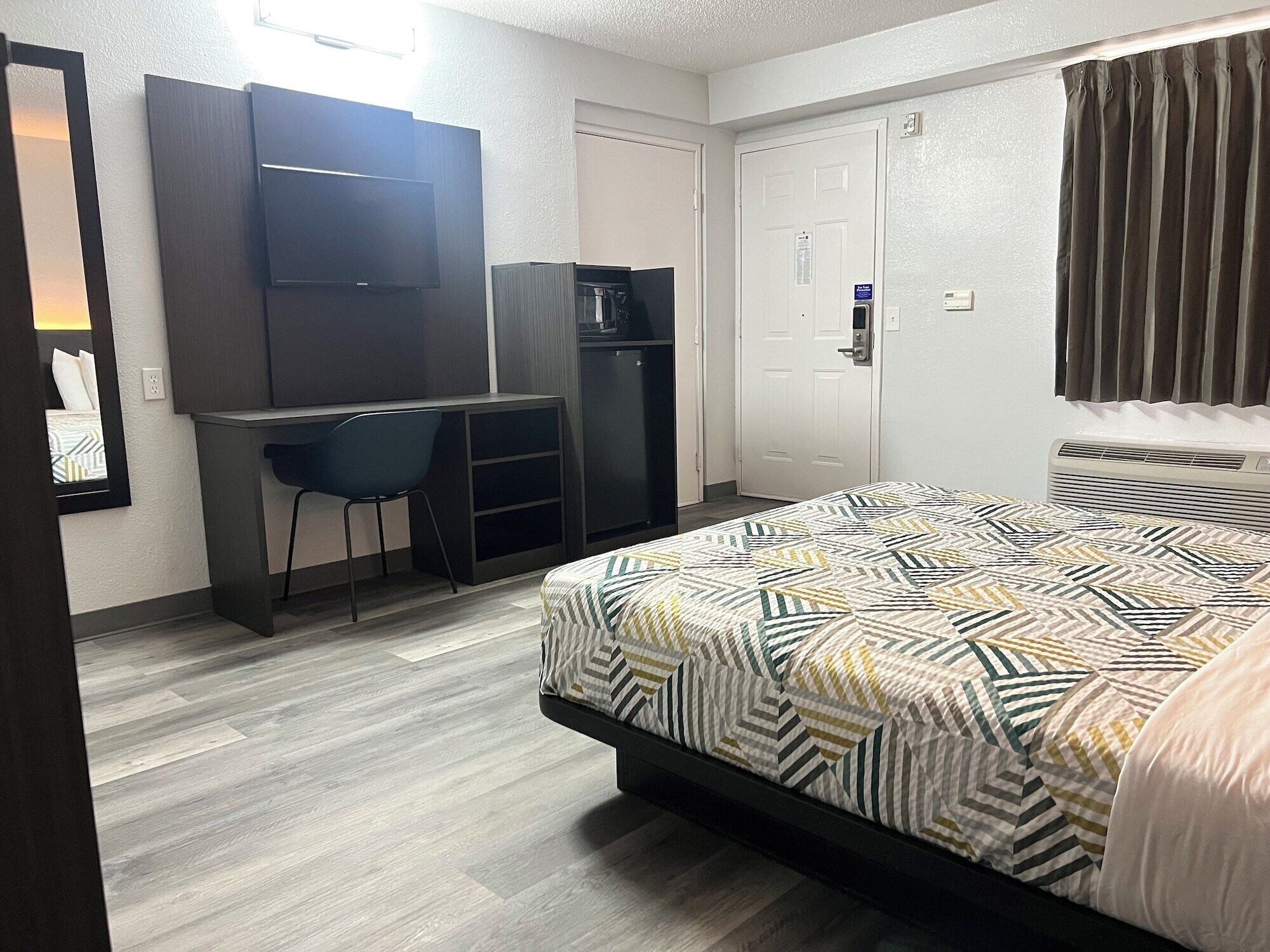 A hotel room with a bed, a desk with a chair, a wardrobe, a door, and a window with curtains.
