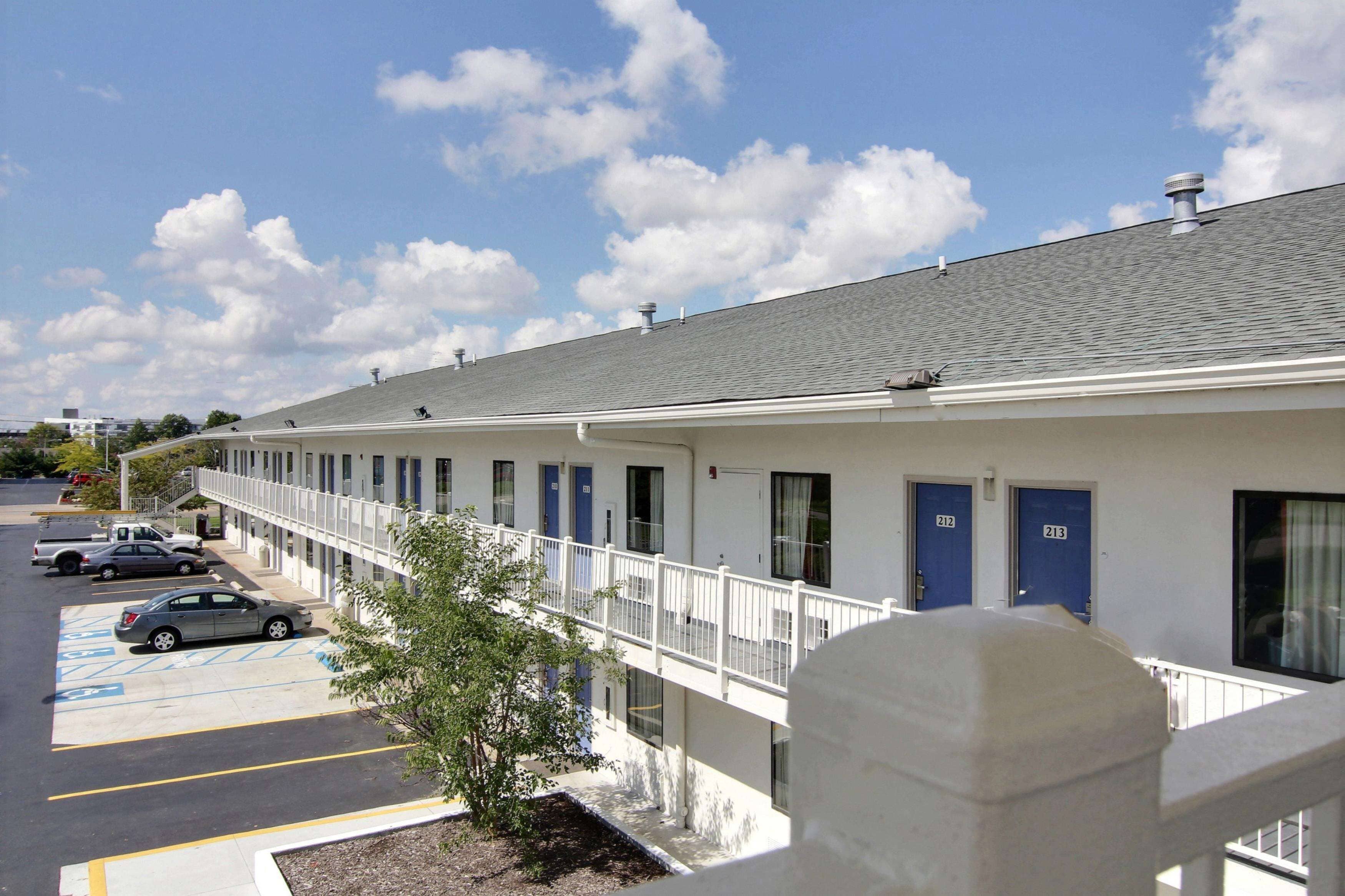 A motel with a row of rooms, a covered parking area, and a small tree in the foreground.