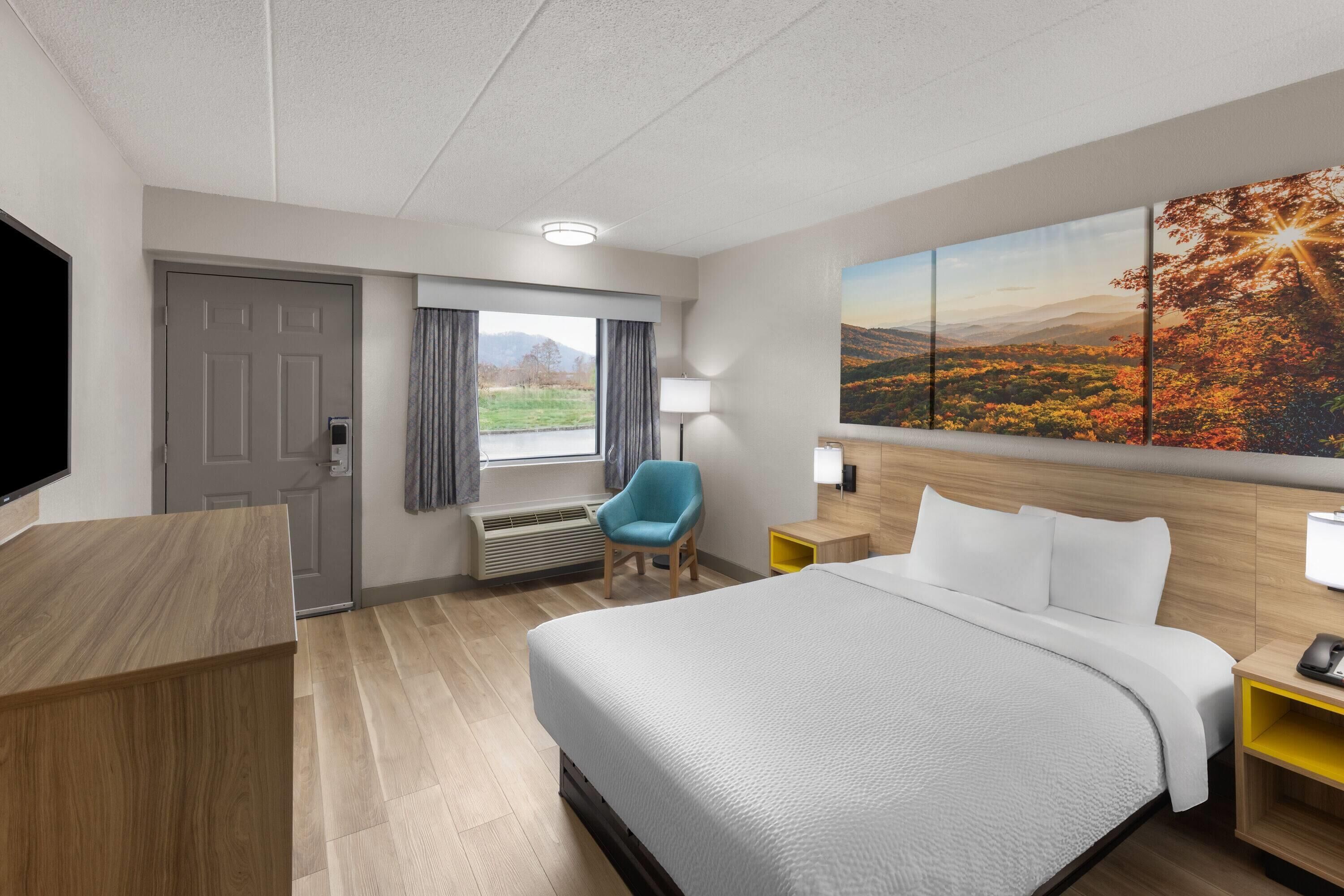 A hotel room with a large bed, a desk, a chair, a TV, a window with curtains, a painting, and a lamp.