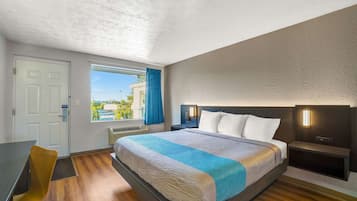 A hotel room with a large bed, a desk, a chair, a window with a view, and a nightstand with a lamp.