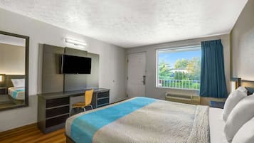 Habitación de hotel con cama, escritorio, silla, televisión y ventana con vista a vegetación.