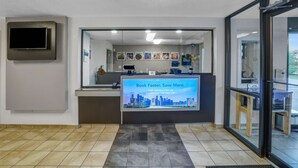 A reception desk with a digital screen displaying a cityscape and promotional text, a flat-screen TV mounted on the wall, and a tiled floor. - Motel 6 Lexington I-75 Hamburg (Lexington)
