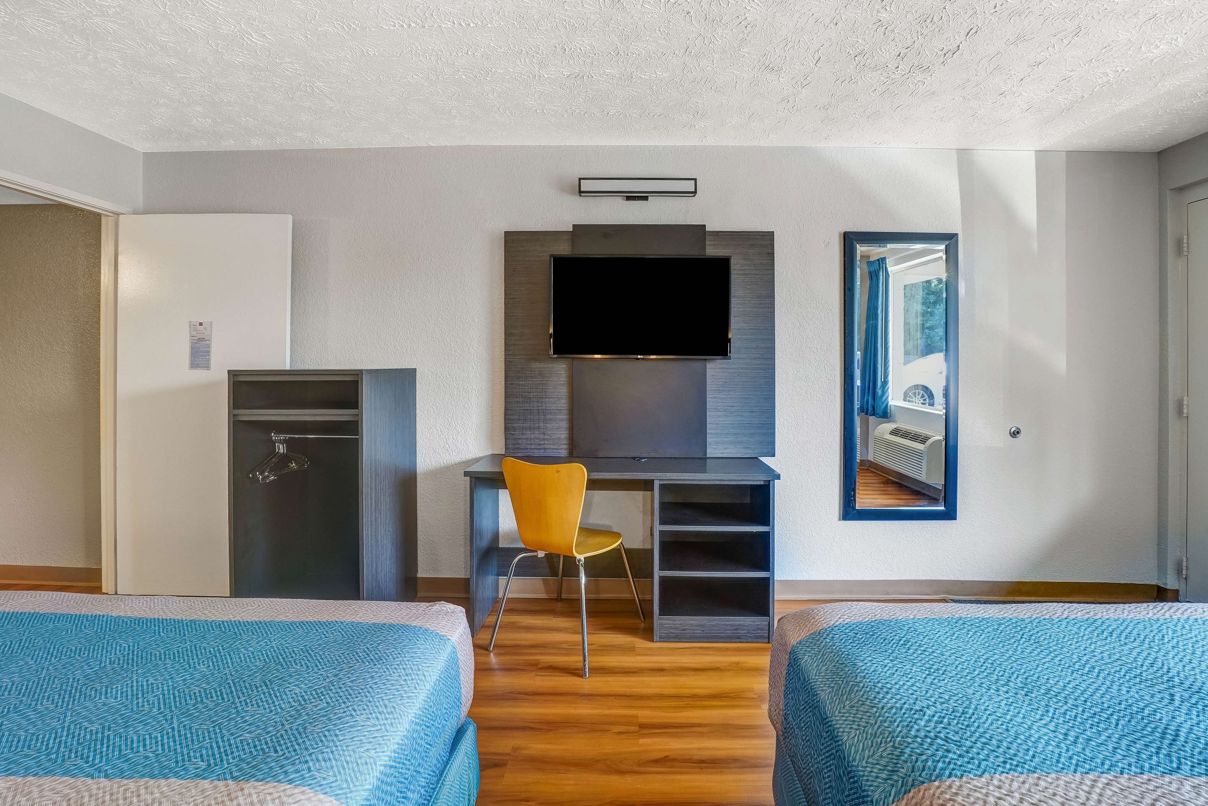 A hotel room with two beds, a desk with a chair, a flat-screen TV mounted above the desk, a wardrobe, and a mirror on the wall.