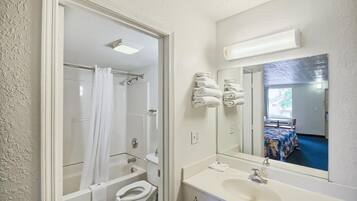 A bathroom with a bathtub, toilet, sink, and a mirror reflecting a bed and window.