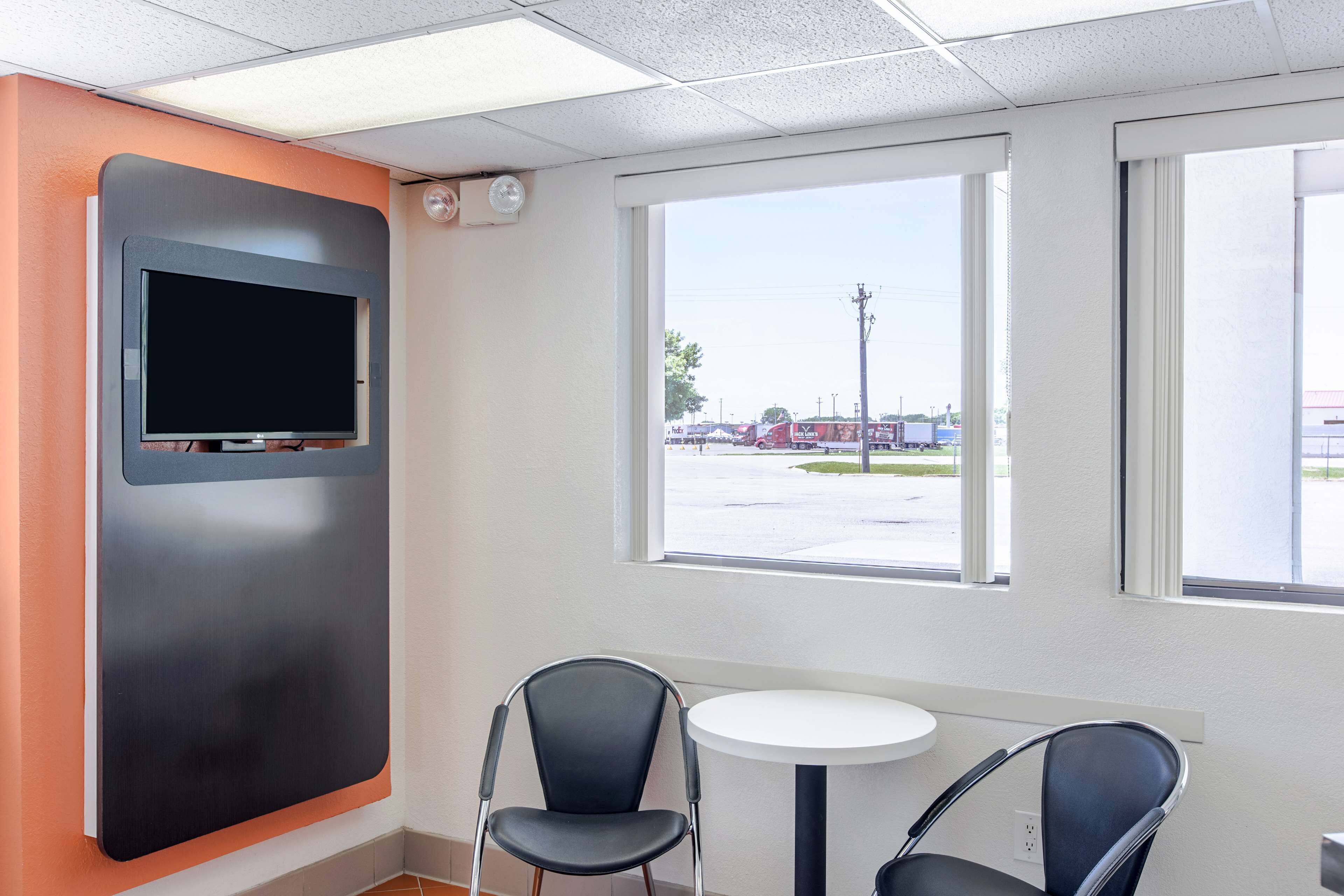 Habitación con una televisión montada en la pared, dos sillas, una mesita y una ventana con vista a un estacionamiento.