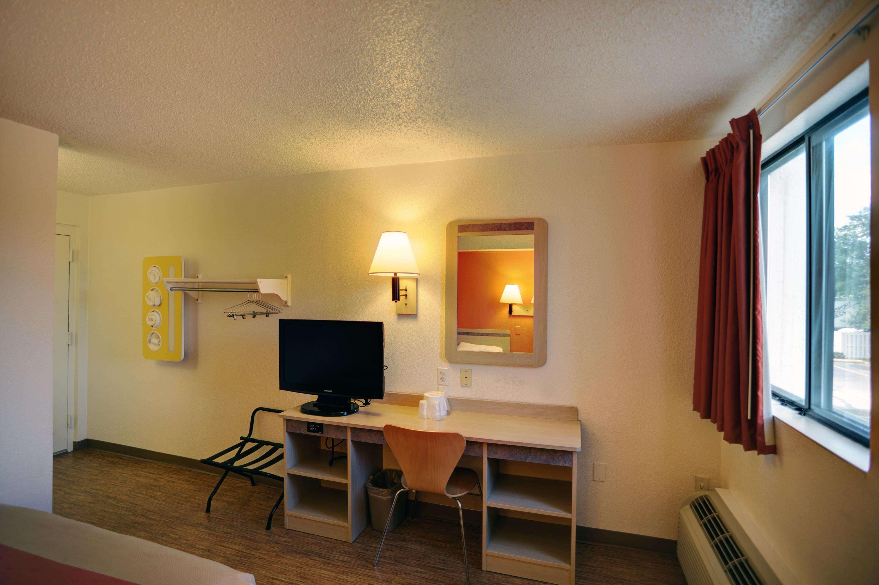 Habitación de hotel con escritorio, silla, televisor, lámpara y ventana con cortinas.