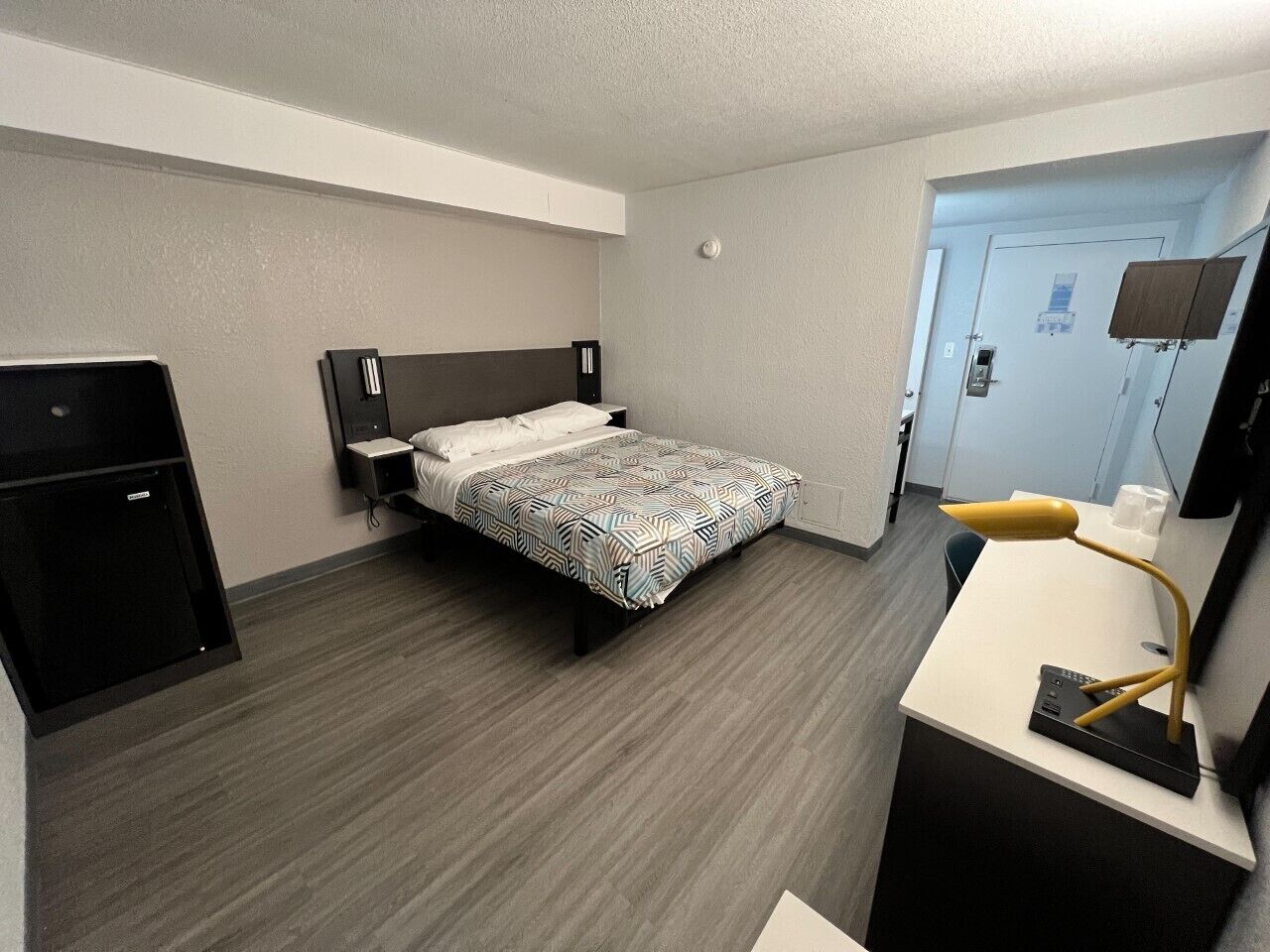 A hotel room with a bed, a desk with a lamp, a small refrigerator, and a television on the wall.