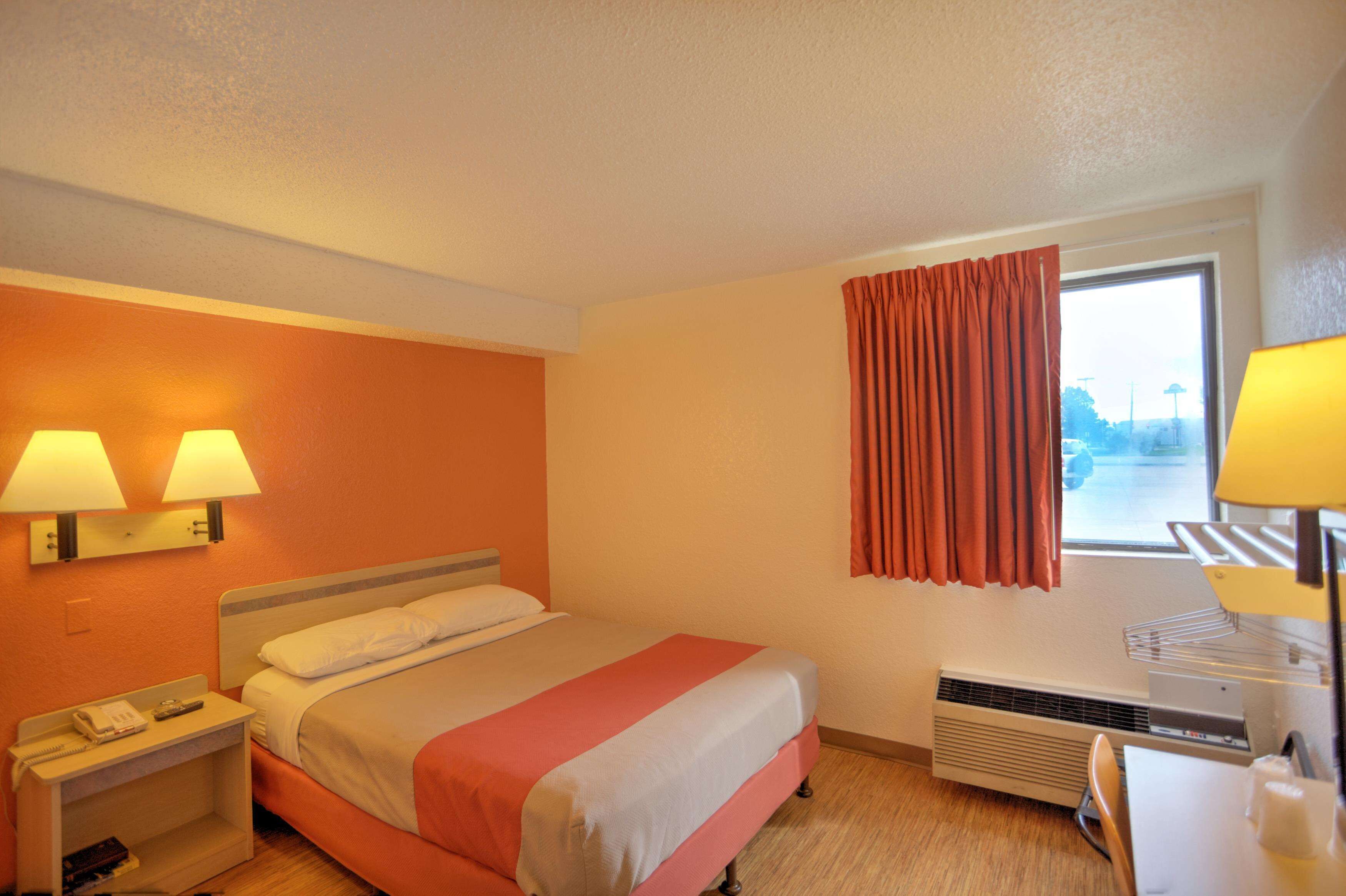 A hotel room with a bed, two bedside lamps, a heater, a window with curtains, and a desk with a lamp.
