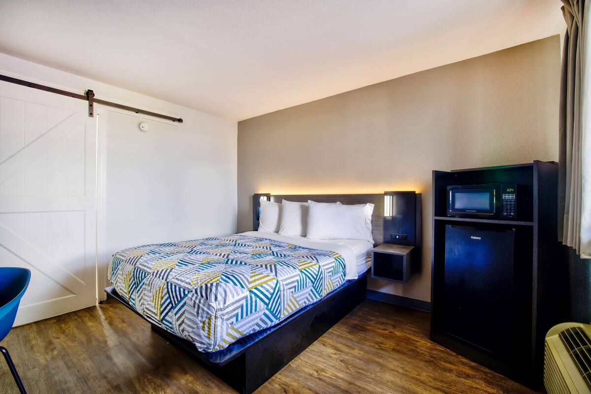 A single bed with a patterned bedspread, a bedside table with a lamp, a microwave, and a refrigerator in a hotel room with wooden flooring and a white door.