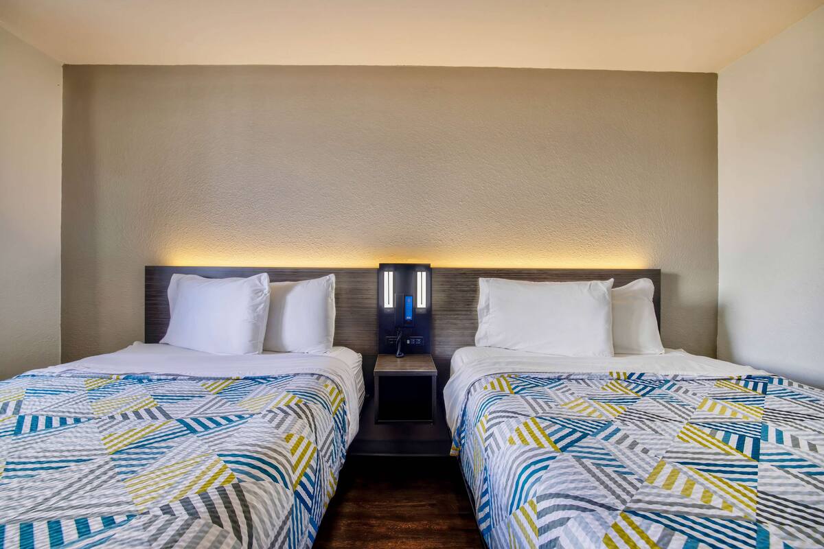 A hotel room with two beds, each with a patterned bedspread, a nightstand with a lamp, and a wall-mounted light fixture.