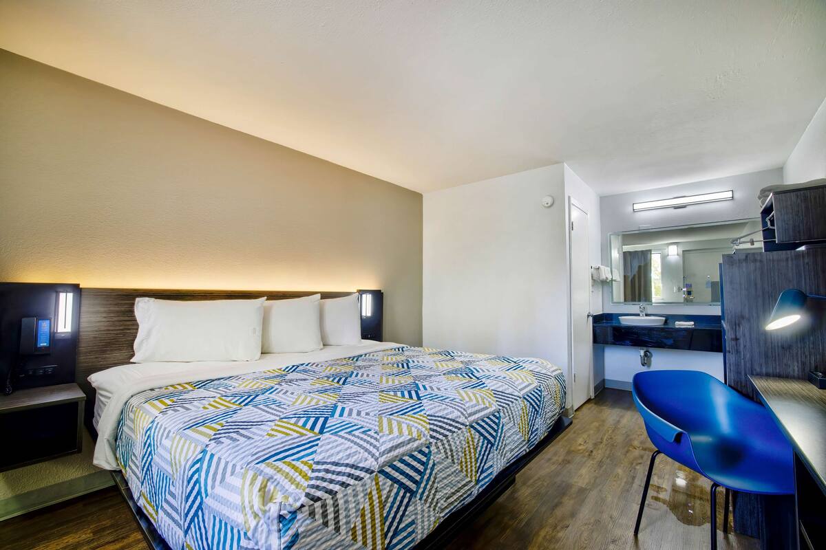 A hotel room with a bed, bedside table, chair, and a bathroom with a sink and mirror.