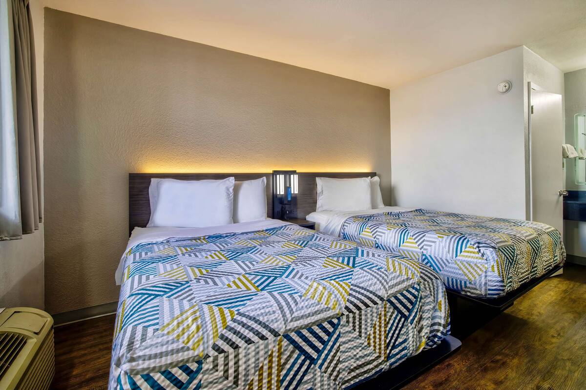 A hotel room with two beds, each with patterned bedding, a wall-mounted light above the beds, and a small bedside table.