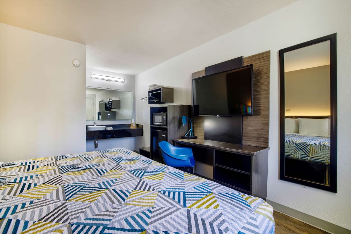 A hotel room with a bed, a desk with a chair, a TV, a mirror, and a bathroom with a sink.