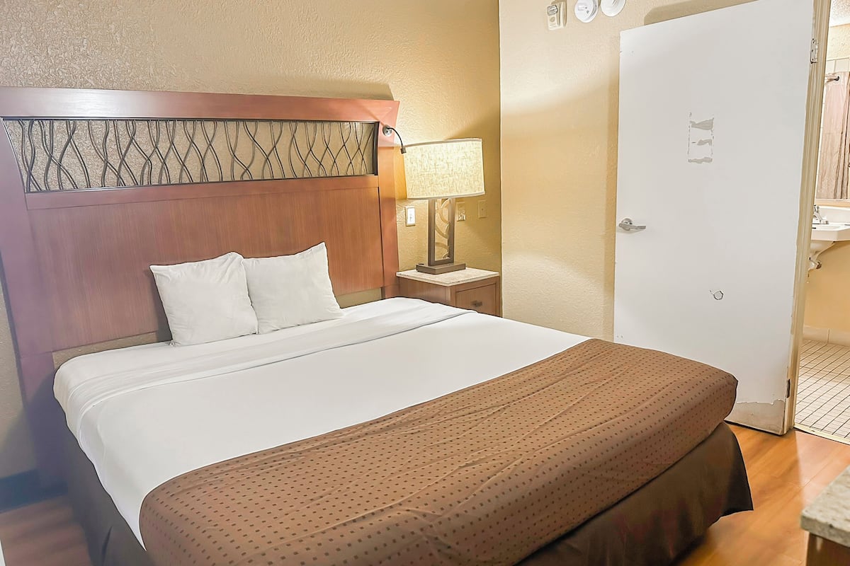 Una habitación de hotel con una cama grande, una mesita de noche con una lámpara y una puerta que da a un baño.