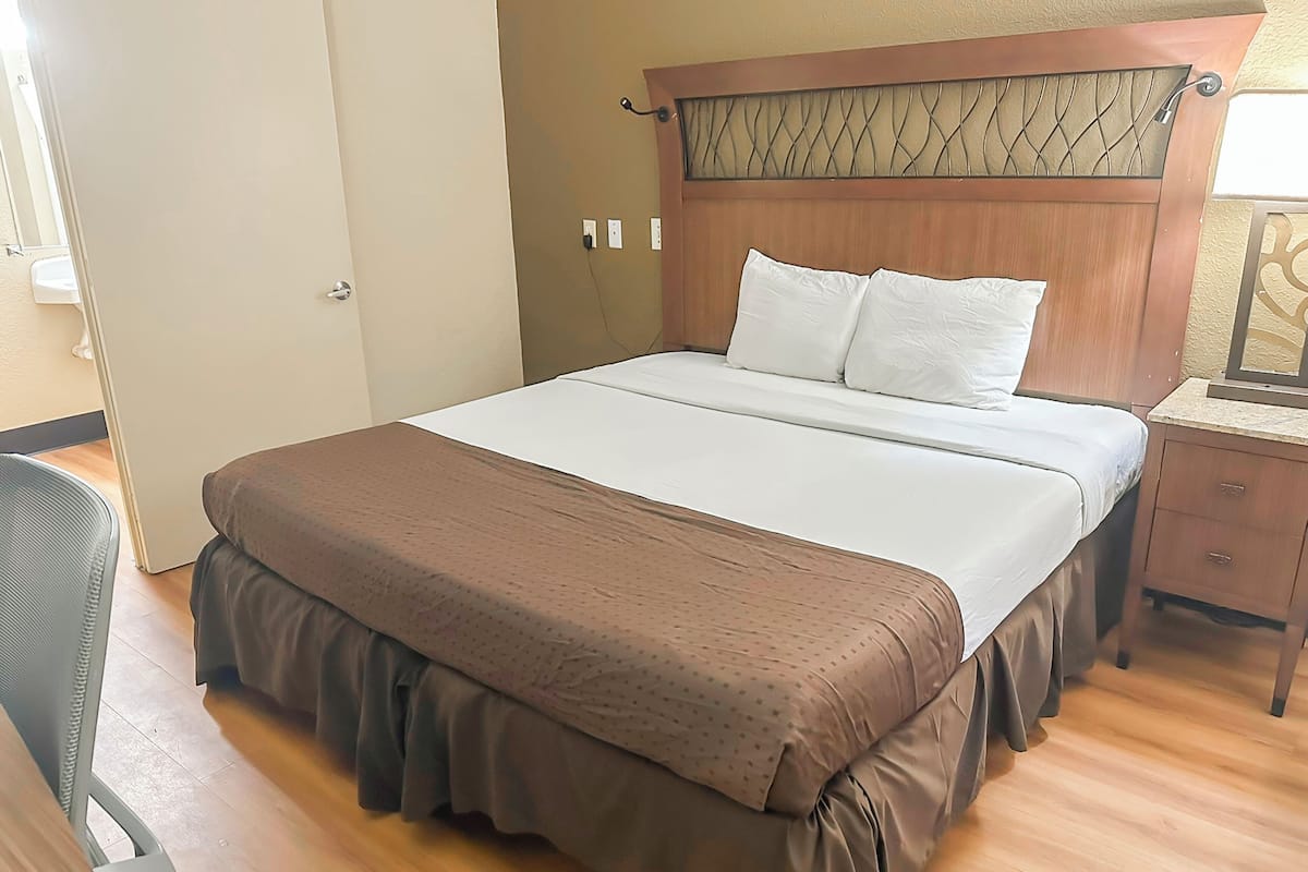 Una cama bien hecha con cabecera de madera, una mesita de noche con lámpara y una silla en una habitación de hotel.