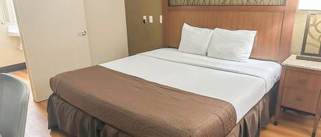 A neatly made bed with a wooden headboard, a bedside table with a lamp, and a chair in a hotel room.