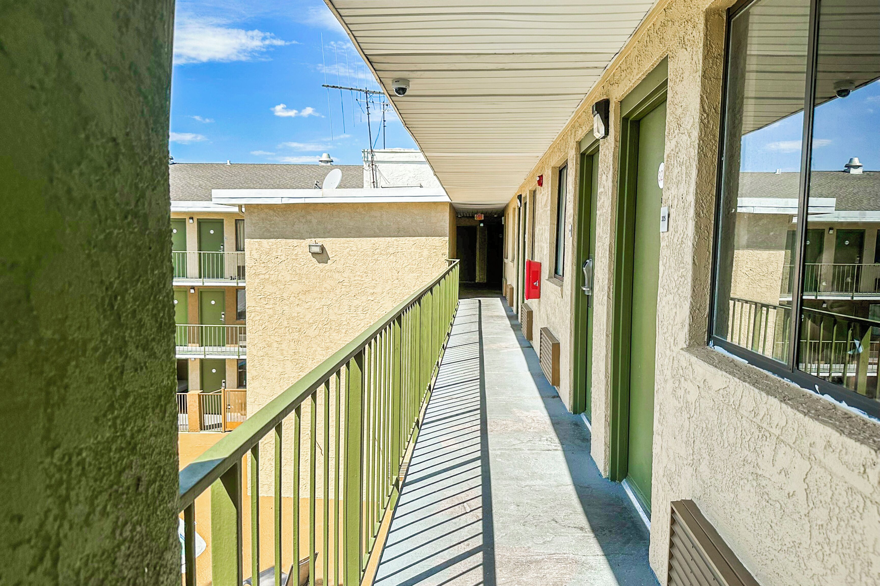 A corridor with a balcony, green doors, and a view of neighboring buildings.