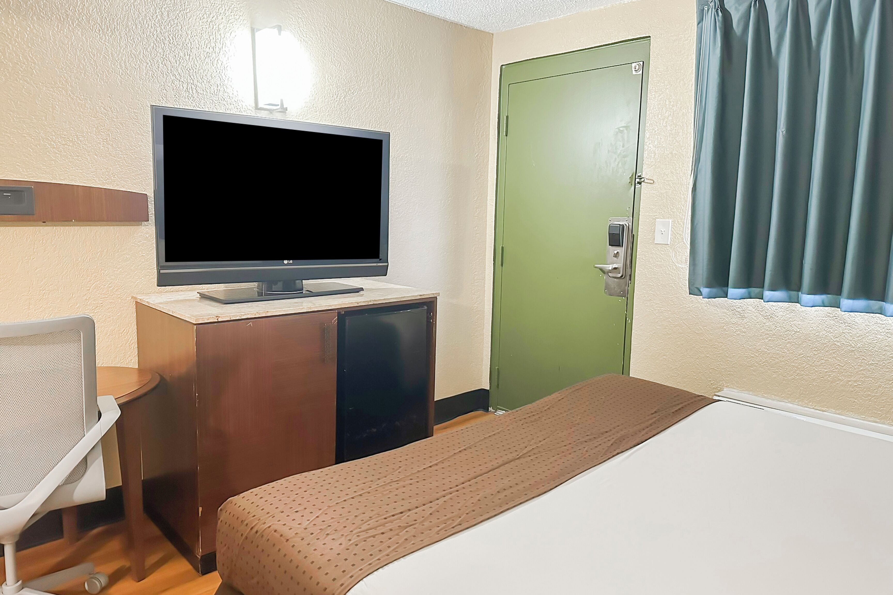 A hotel room with a bed, a desk with a television, a small refrigerator, a chair, and a green door.