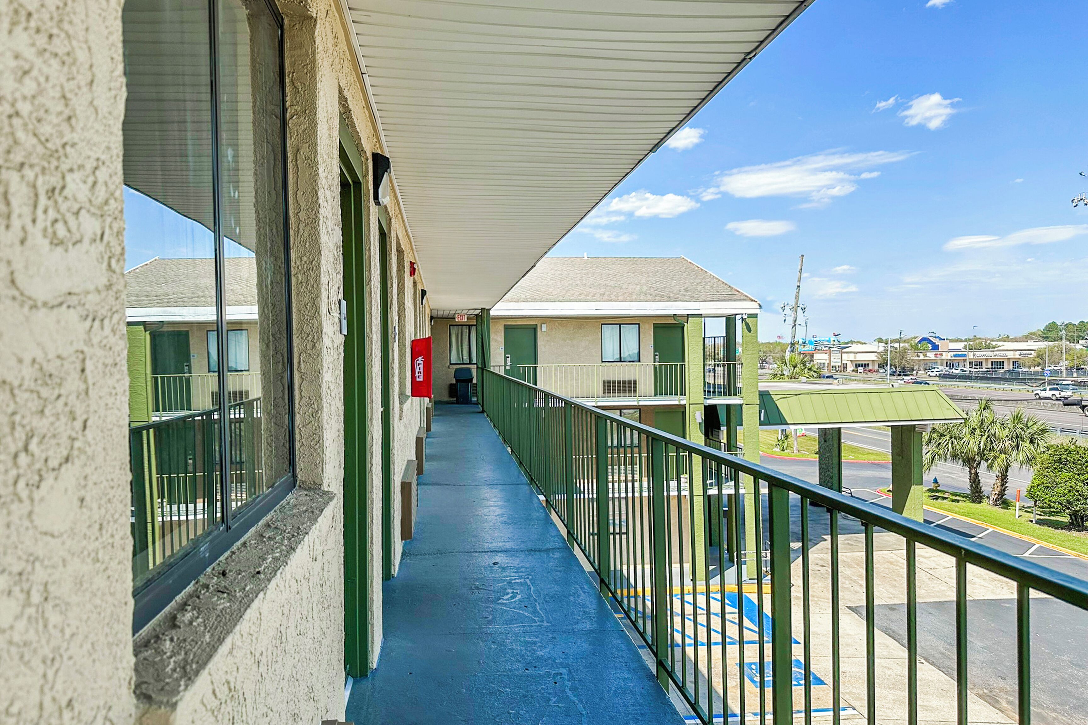 A corridor with a blue floor, green railings, and a view of a parking lot and buildings in the distance.