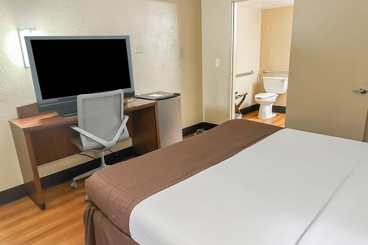 Habitación de hotel con cama, escritorio con computadora y silla, televisión, y un baño visible a través de una puerta abierta.