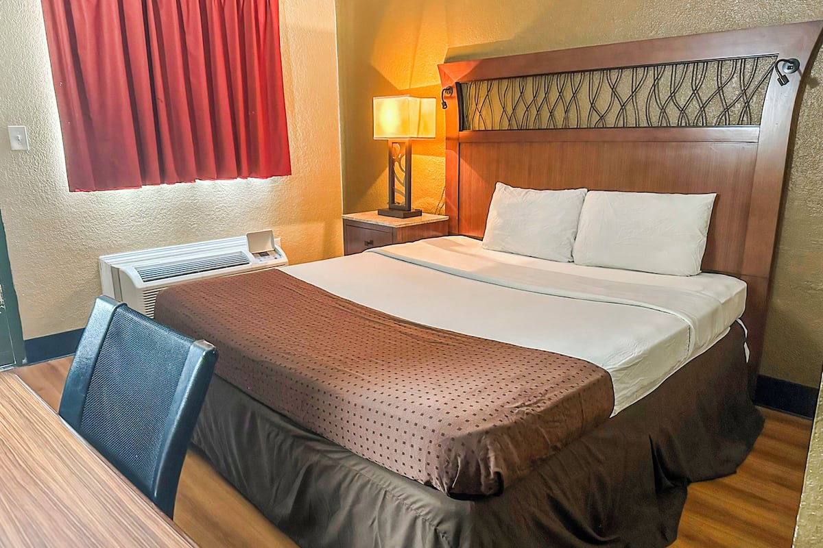 Habitación de hotel con cama, escritorio, silla, lámpara y ventana con cortinas rojas.