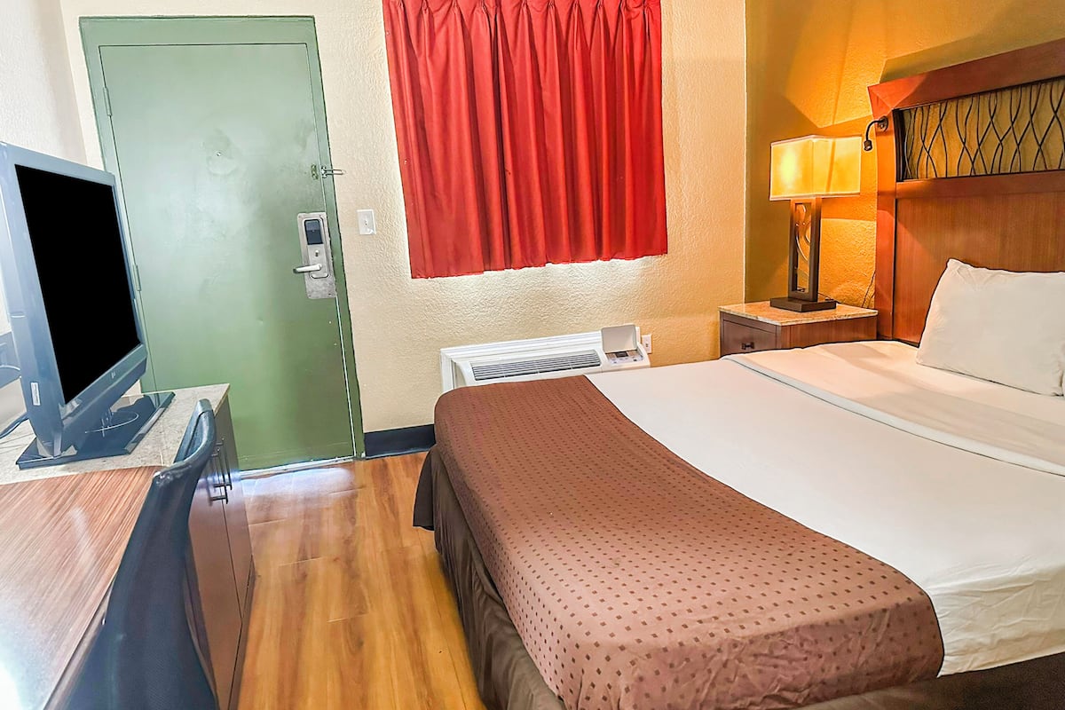 Habitación de hotel con cama, televisor, calefacción, ventana con cortinas rojas y mesita de noche con lámpara.