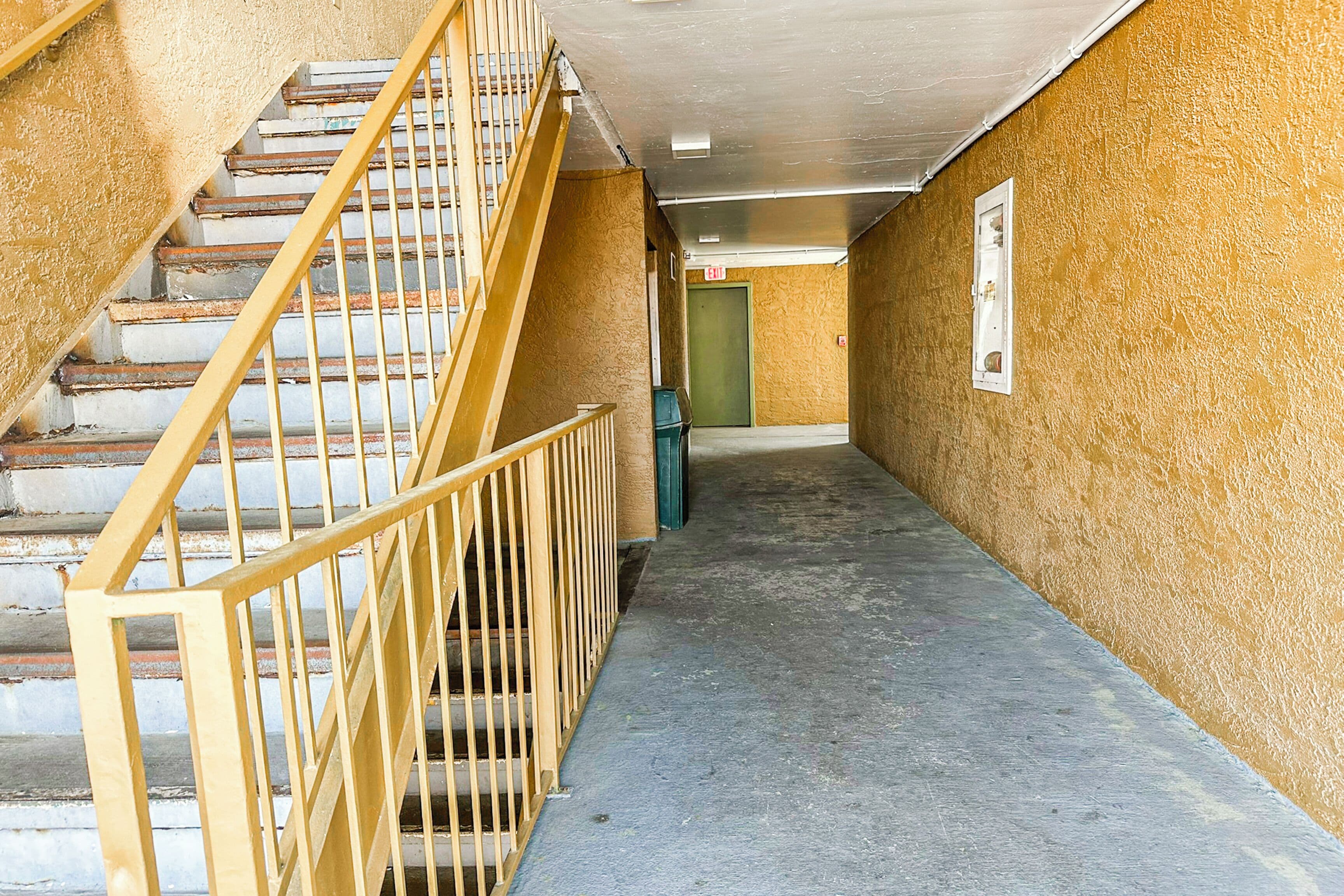 A narrow hallway with a staircase on the left, a green door at the end, and a trash bin in the middle.