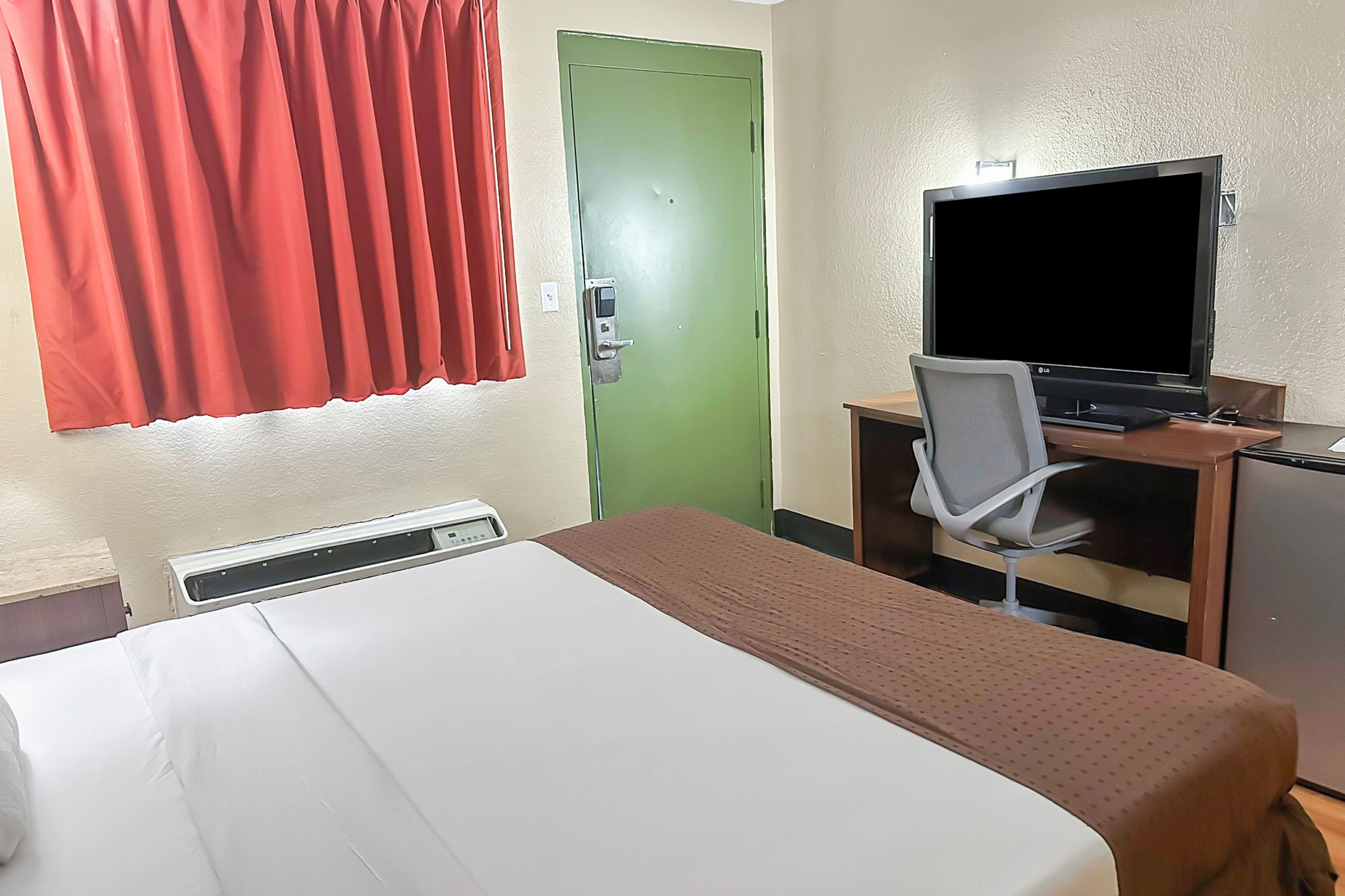 A hotel room with a bed, a desk with a television, a chair, and a green door.