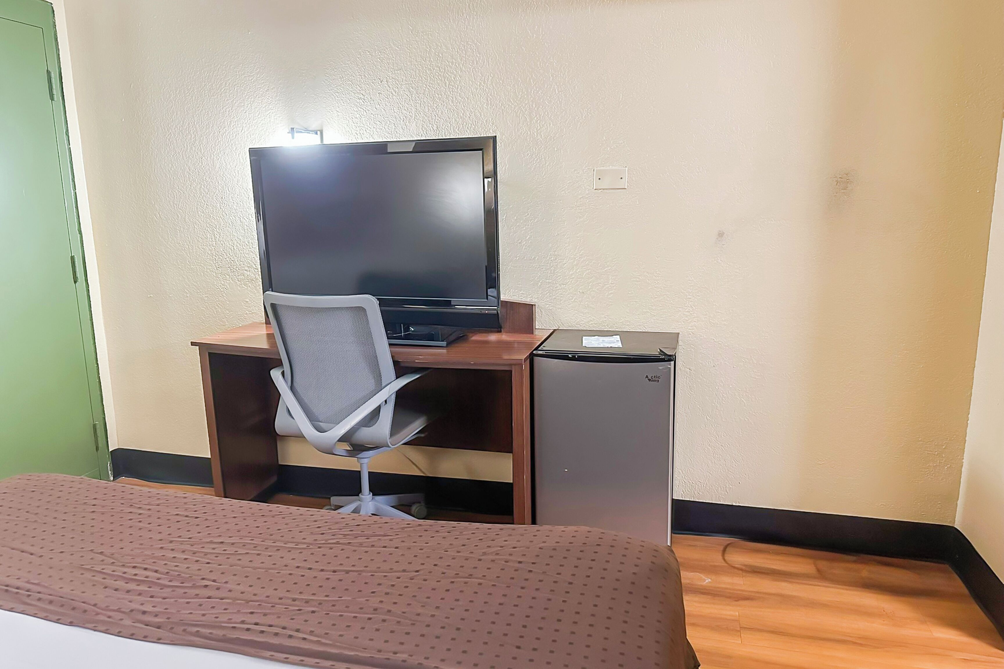 Habitación de hotel con cama, escritorio con computadora y refrigerador pequeño.