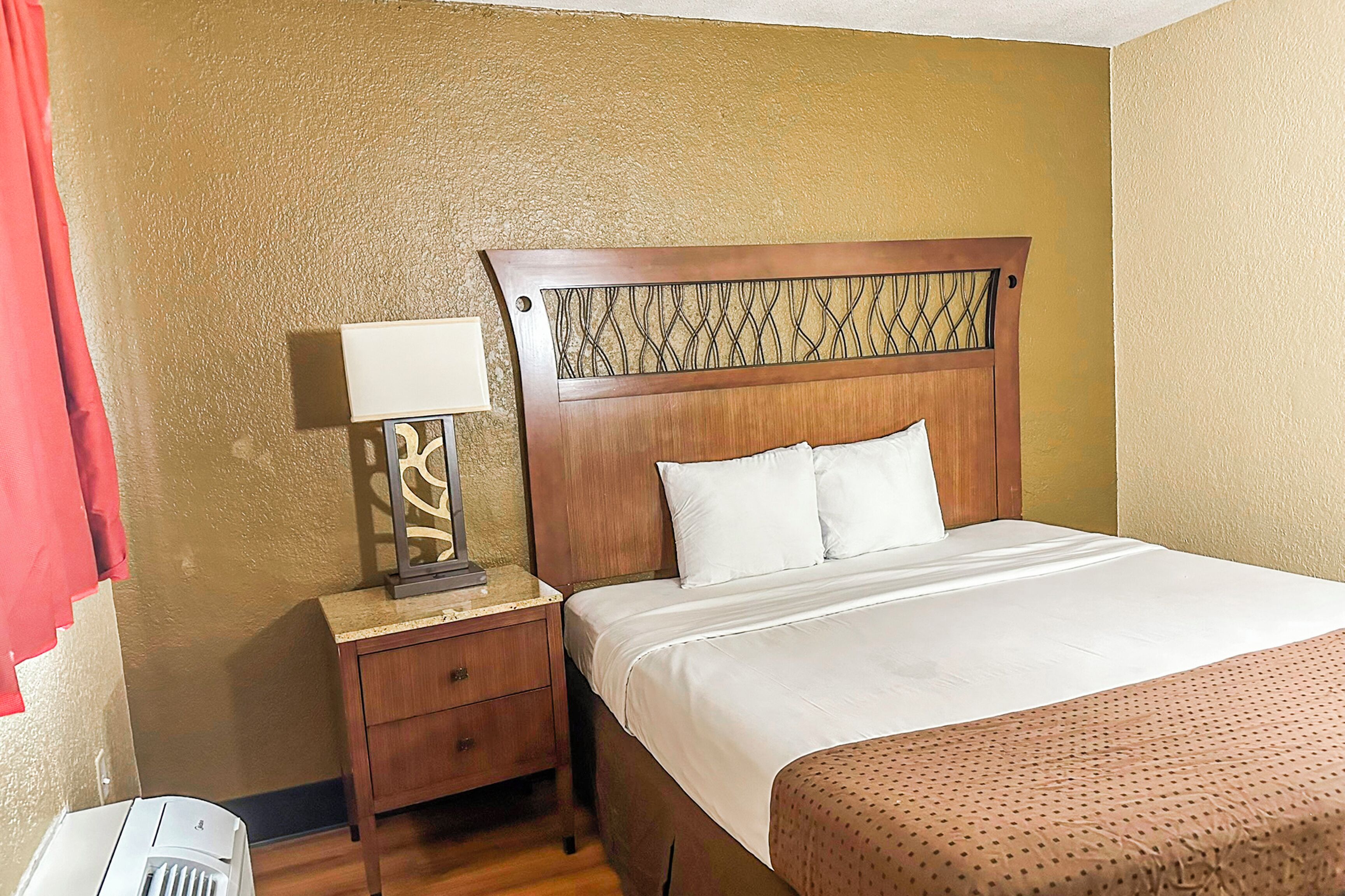 A hotel room with a wooden headboard, a bedside table with a lamp, a bed with white bedding, and a red curtain.