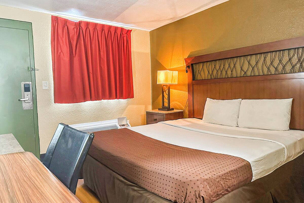 Habitación de hotel con cama, escritorio, silla, mesita de noche con lámpara y cortina roja.