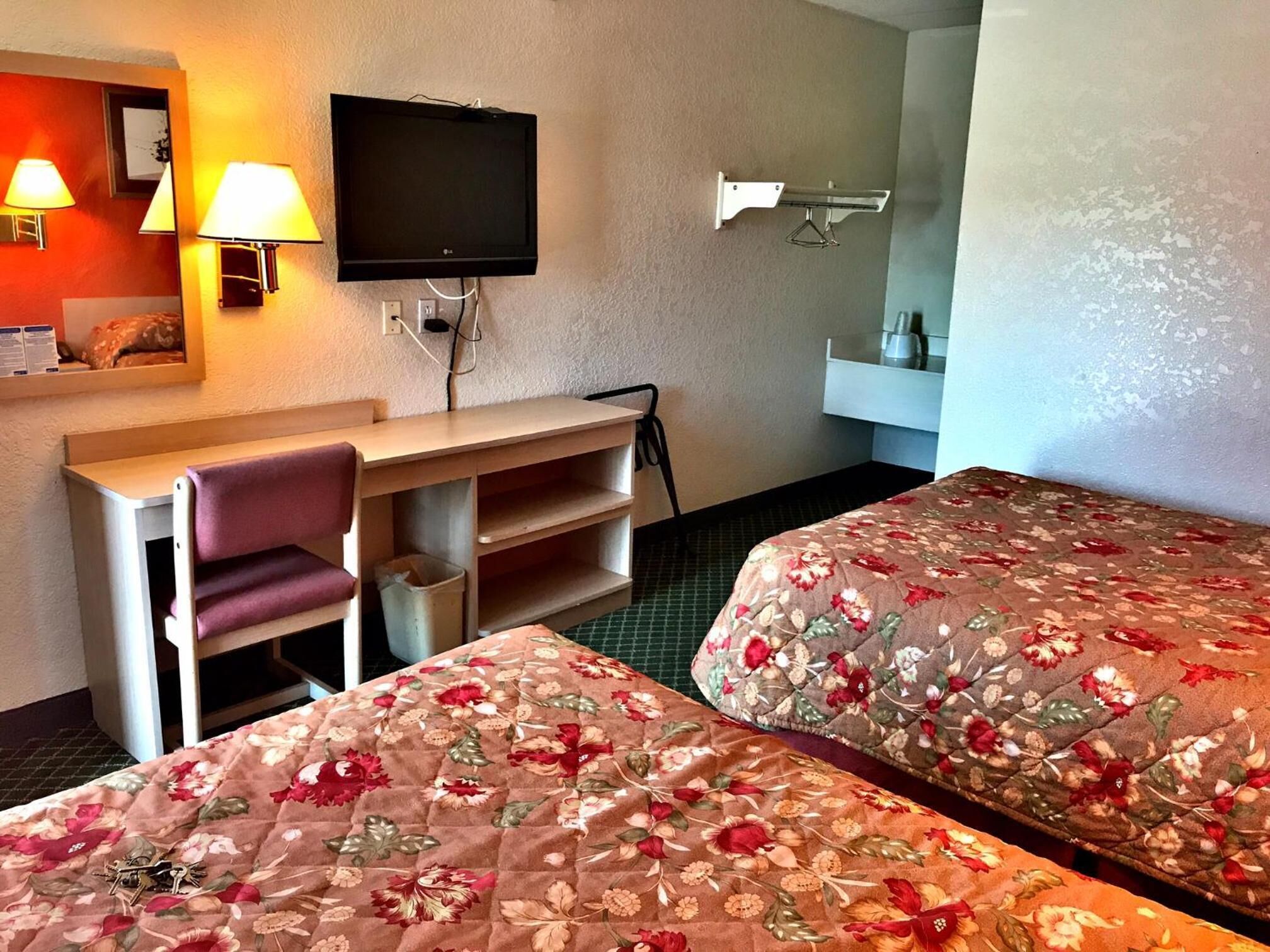 A hotel room with a desk, two beds with floral bedding, a TV, and a mirror.