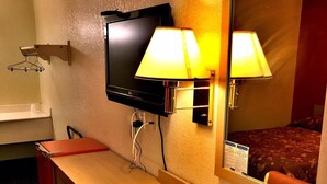A hotel room with a wooden desk, a television mounted on the wall, two bedside lamps, and a mirror reflecting the bed. - QC Stay Inn (Moline)