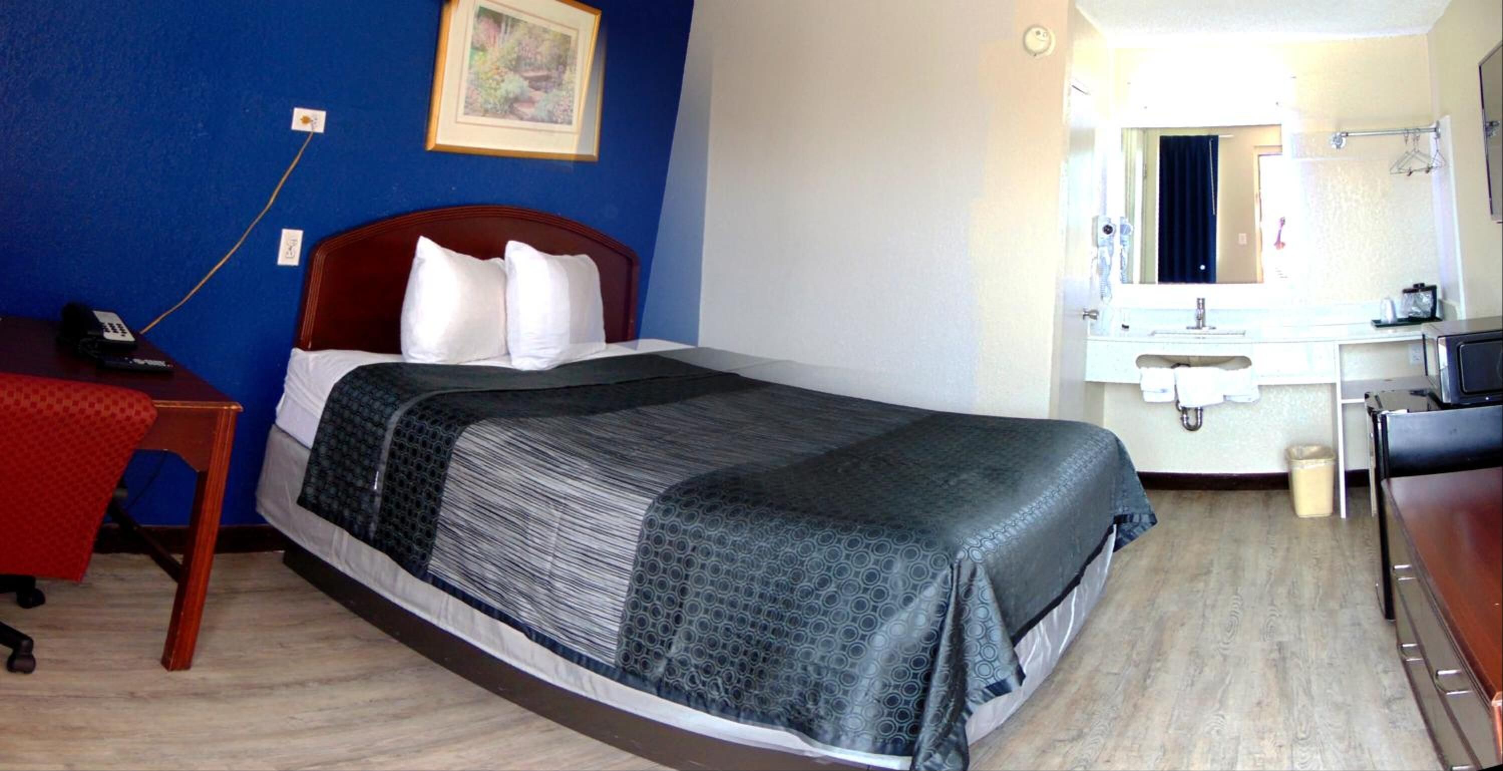 Una camera d'albergo con un letto, una scrivania con il telefono, un bagno con lavandino e specchio, e un piccolo cestino.