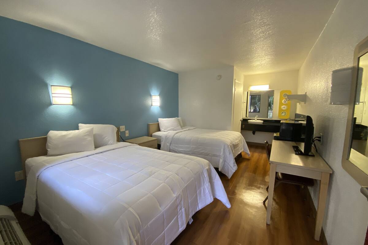 A hotel room with two beds, a desk, a mirror, and a sink.