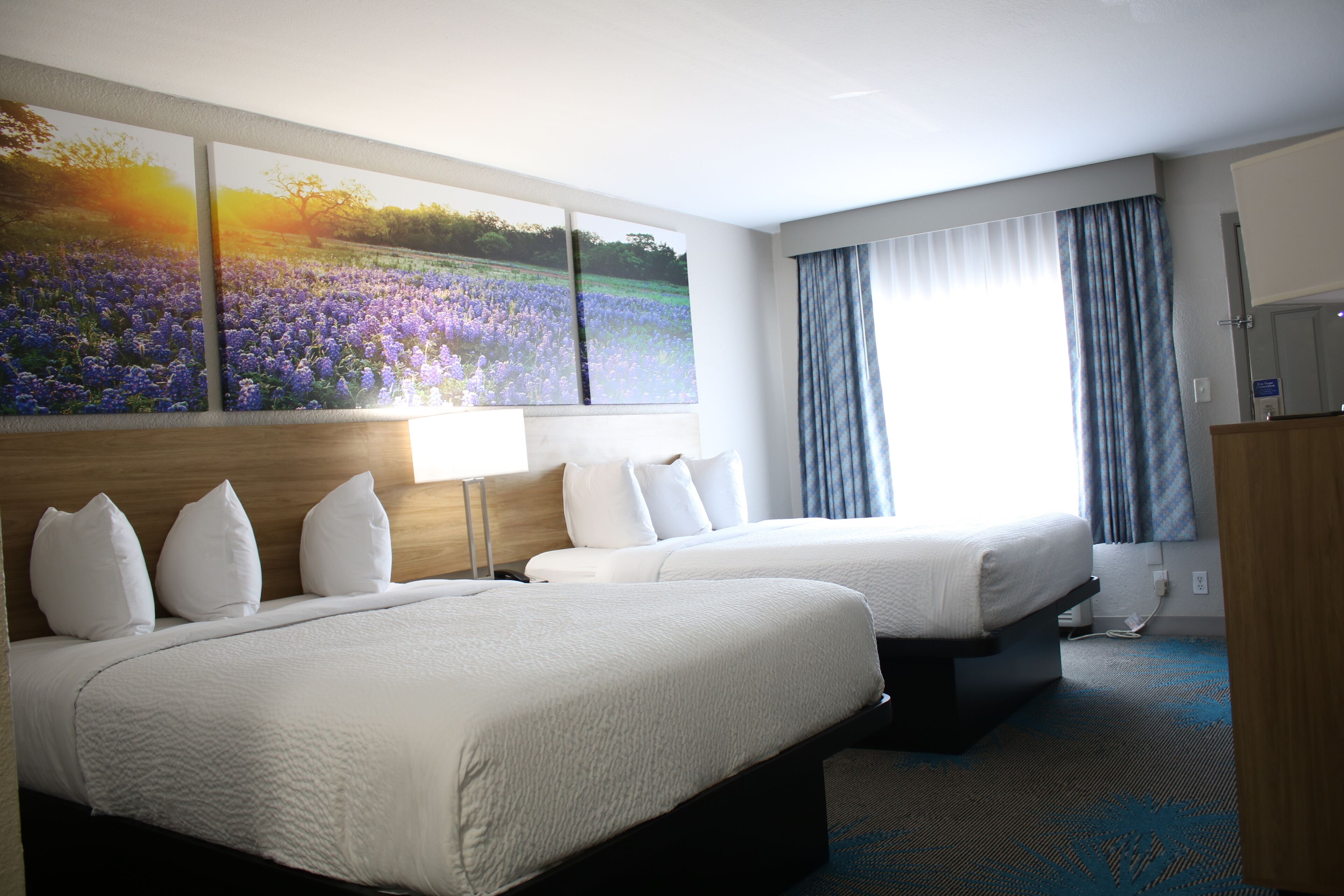 A hotel room with two beds, a large window with curtains, and a painting of a field.
