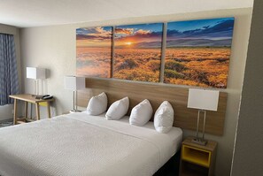 Une chambre d’hôtel avec un grand lit, un bureau, deux lampes de chevet et un tableau représentant un paysage au coucher du soleil, accroché au mur.