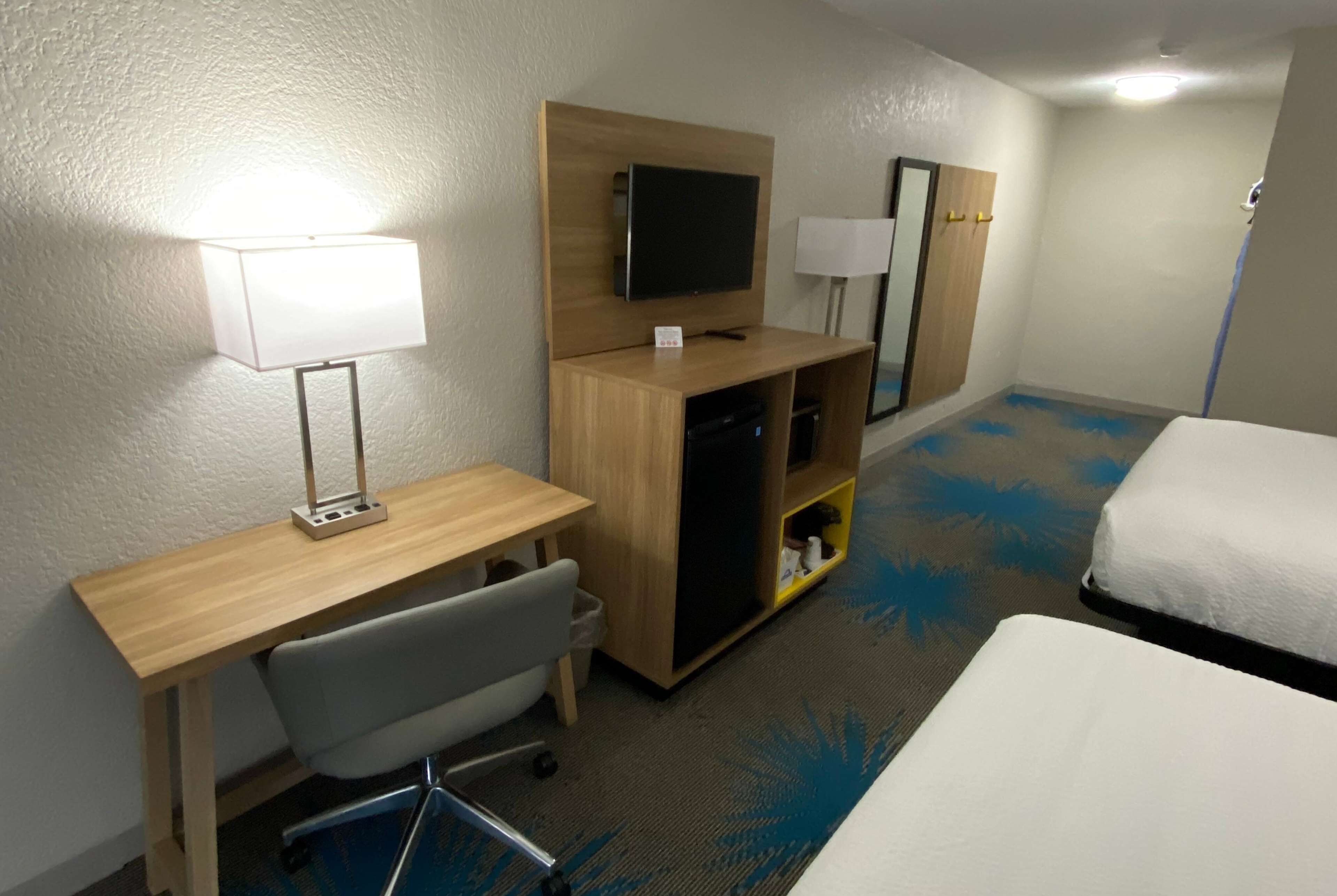 A hotel room with a bed, a desk with a chair, a television, a small shelf, and a lamp.