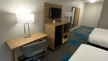 A hotel room with a bed, a desk with a chair, a television, a small shelf, and a lamp.