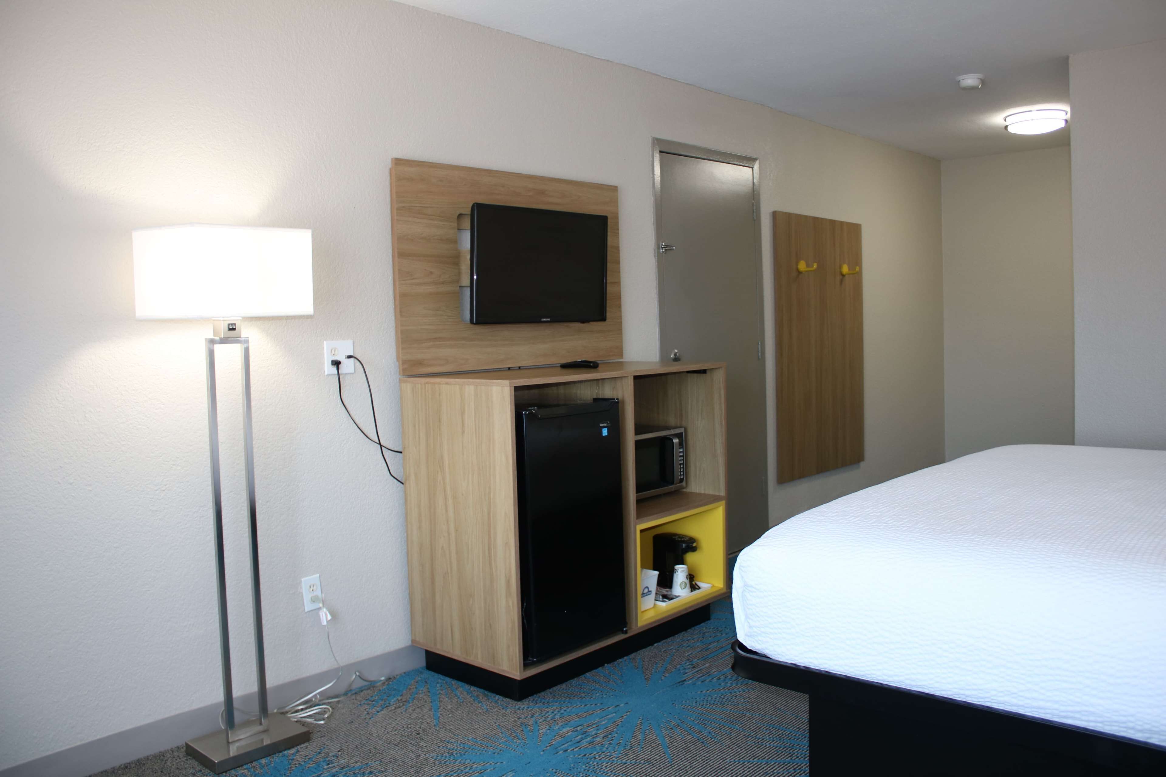 A hotel room with a bed, a TV mounted on the wall, a minibar, a desk, and a lamp.