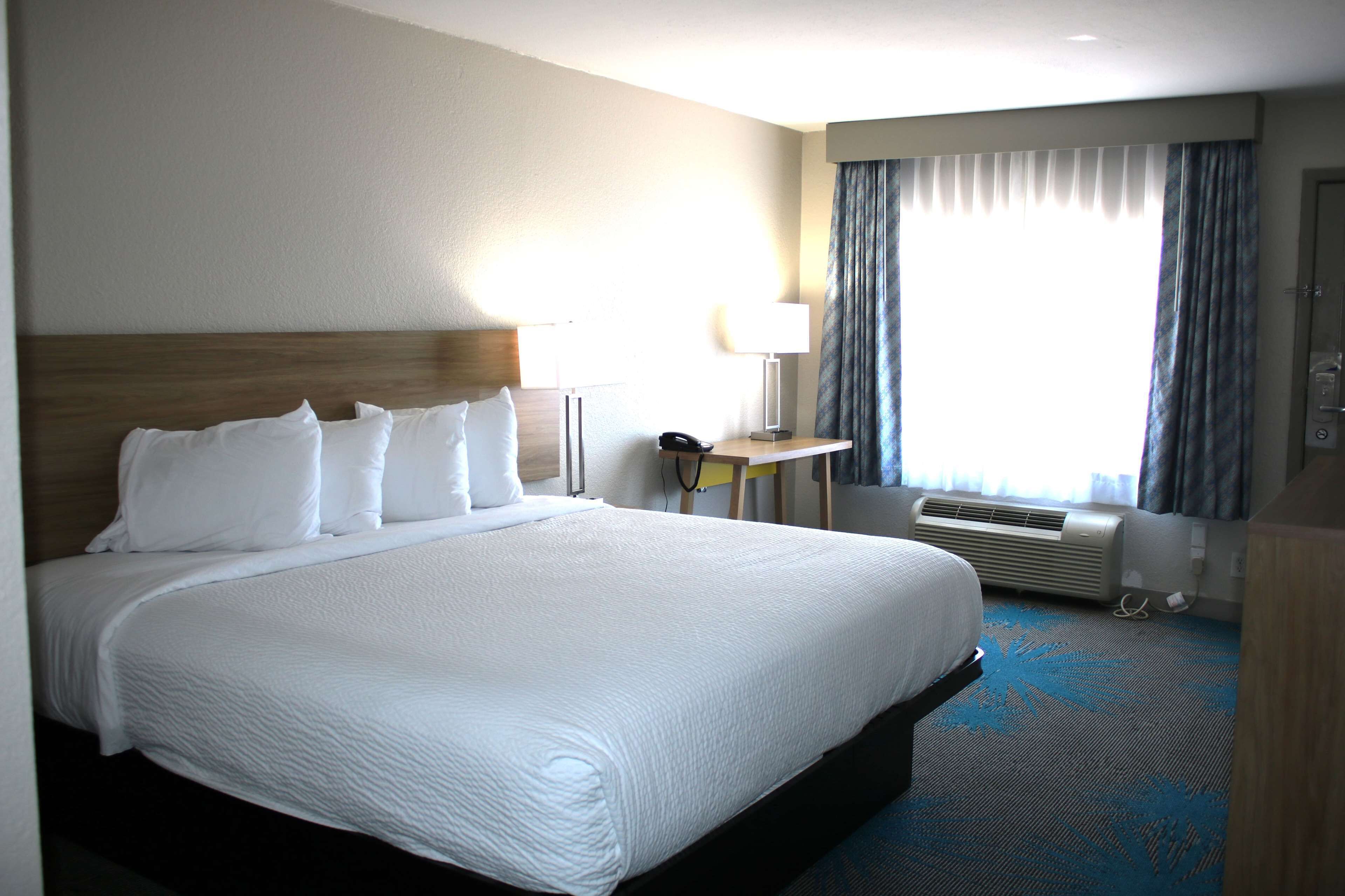 A hotel room with a large bed, a bedside table with a lamp, a desk, and a window with curtains.