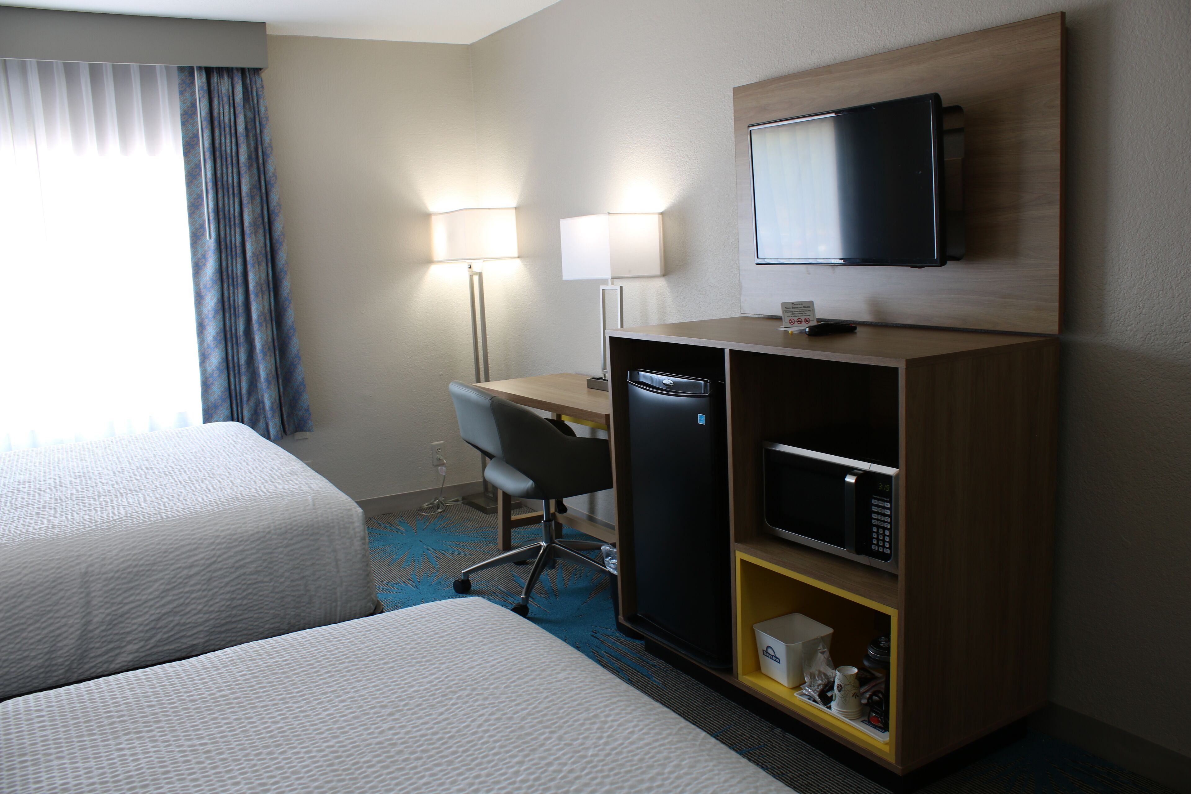 Habitación de hotel con dos camas, un escritorio con silla, televisor, microondas y un estante con objetos.