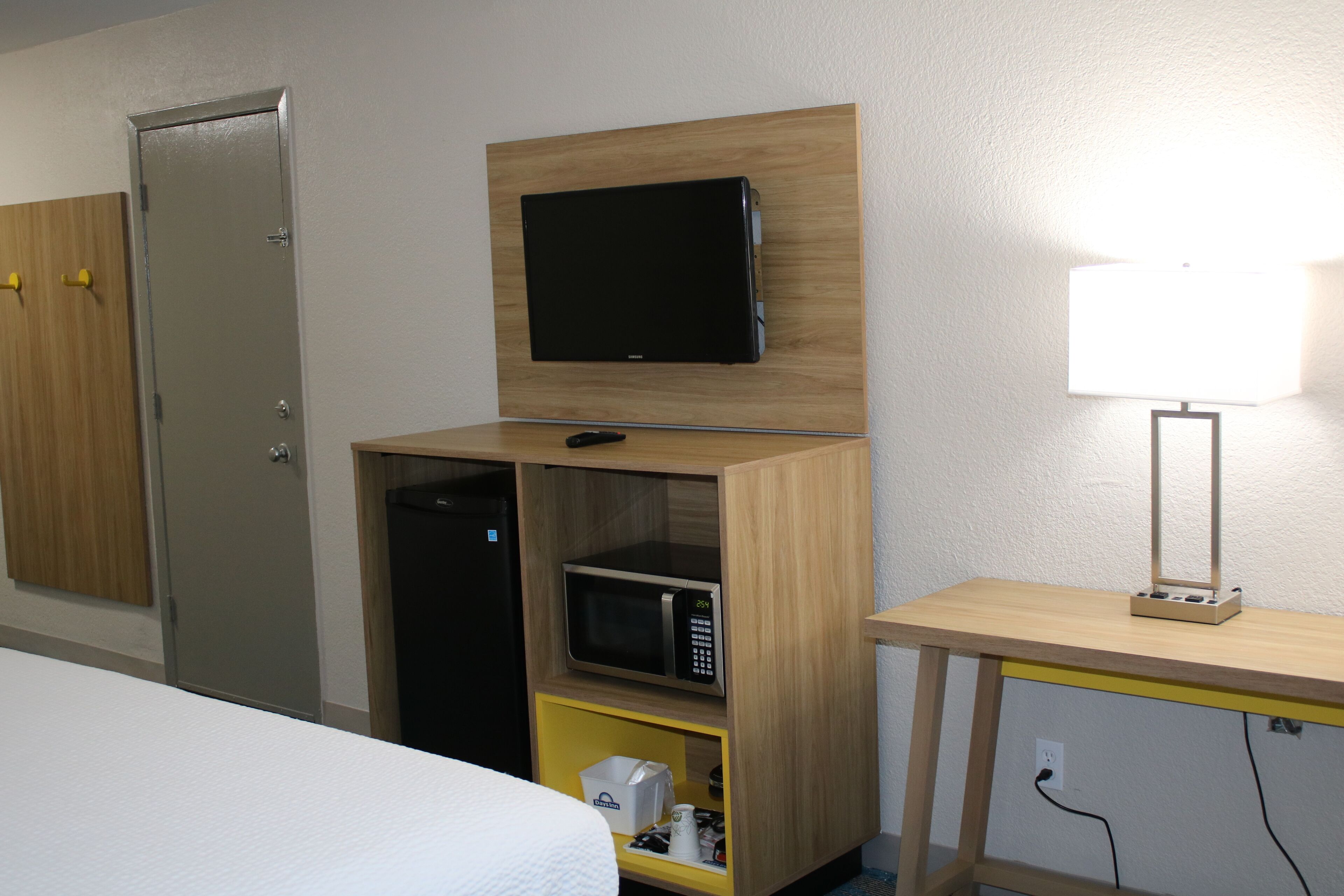 A hotel room with a bed, a TV mounted on a wooden shelf, a microwave, and a desk with a lamp.