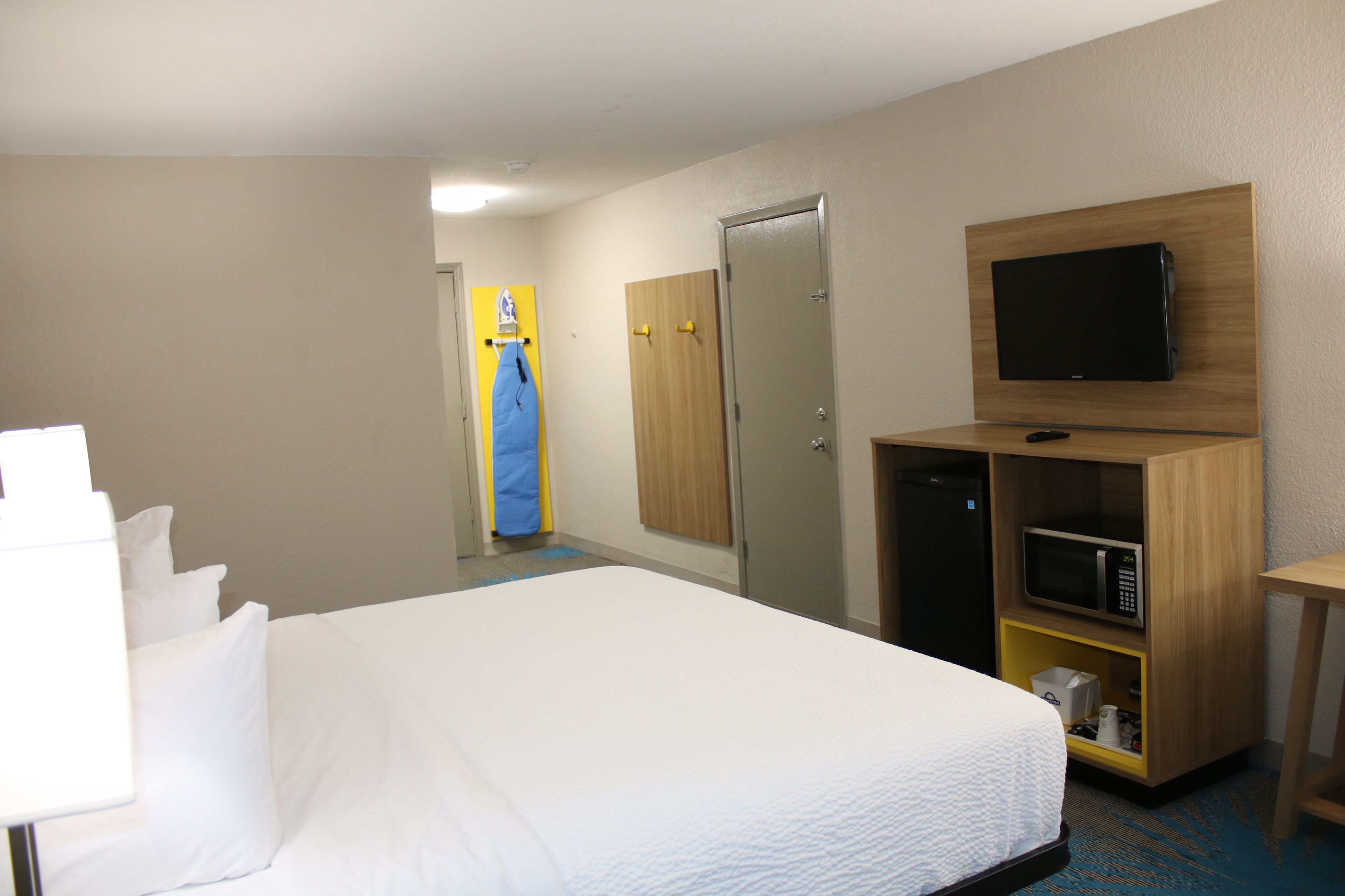 Habitación de hotel con cama, televisor montado en la pared, microondas en un mueble de madera y puerta que da a otra habitación.