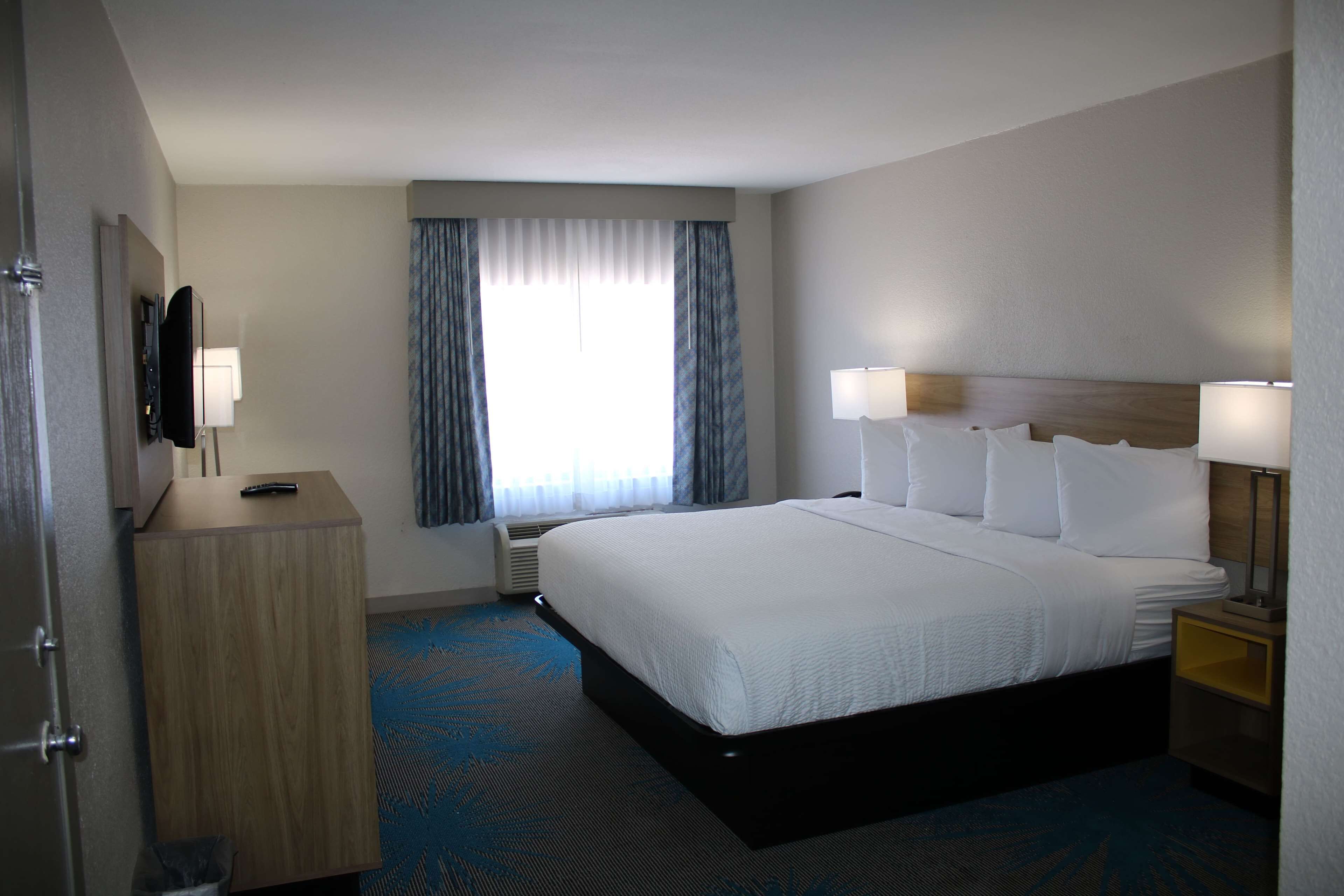 A hotel room with a large bed, a bedside table with a lamp, a TV mounted on the wall, and a window with curtains.