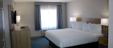 A hotel room with a large bed, a bedside table with a lamp, a TV mounted on the wall, and a window with curtains.
