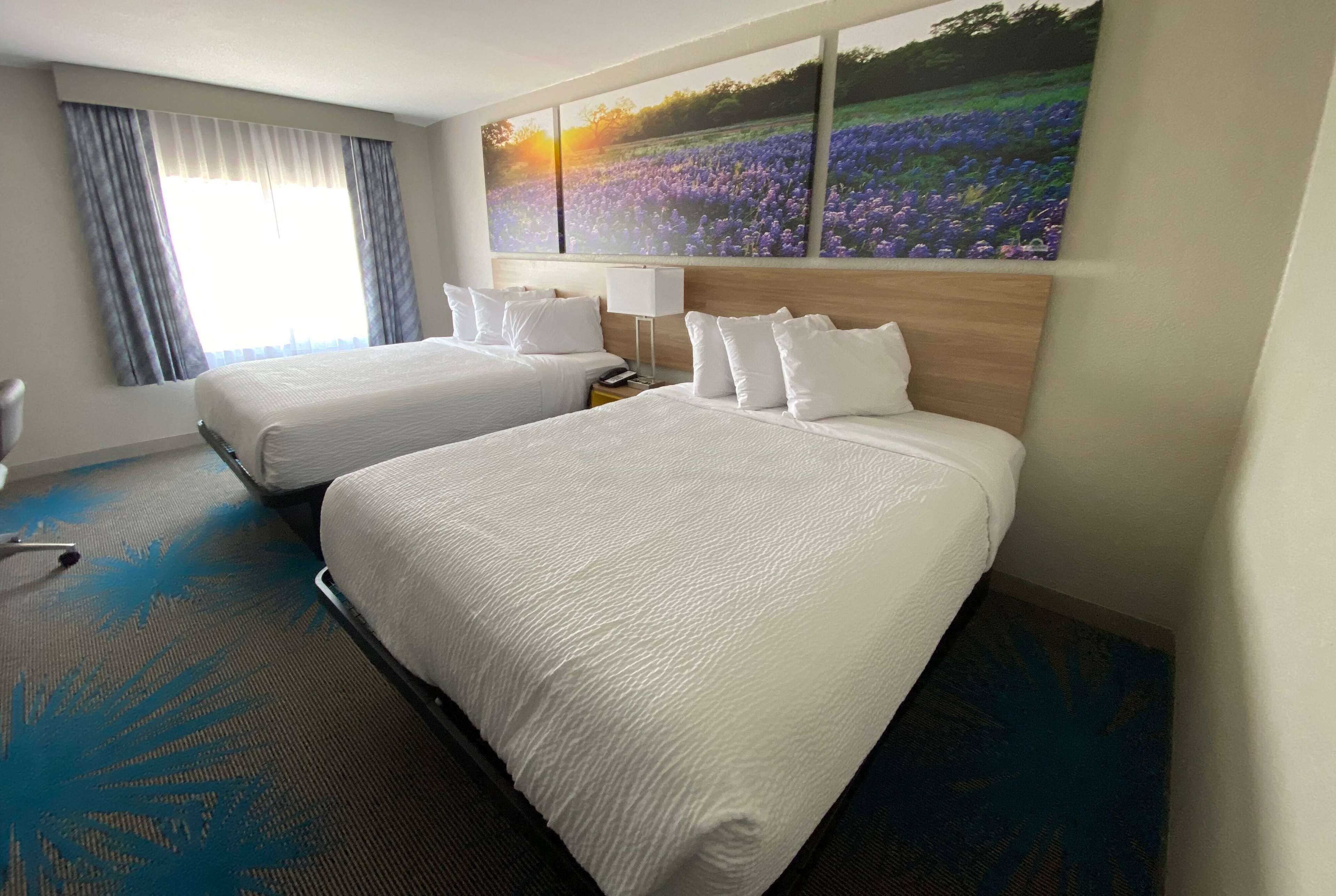 Une chambre d’hôtel avec deux lits, une grande fenêtre avec des rideaux, un tableau représentant un champ et une moquette bleue.