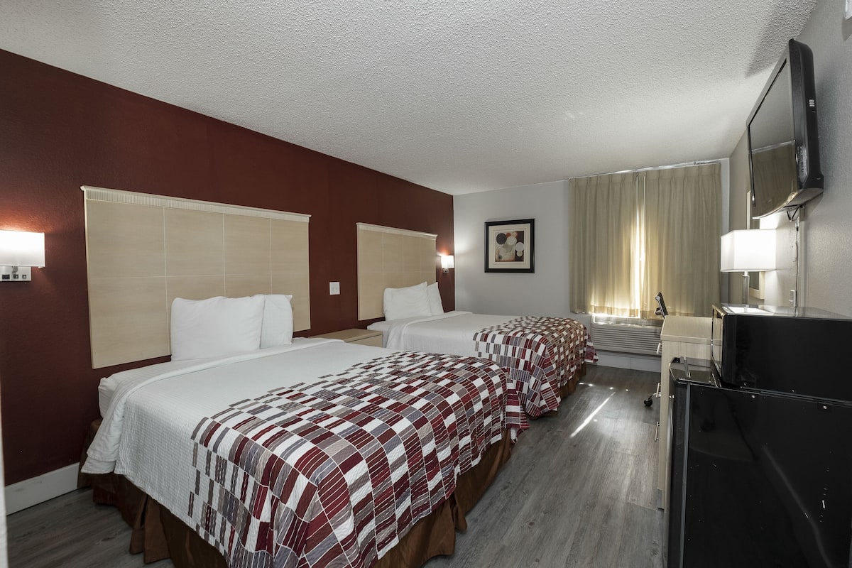 A hotel room with two beds, a television, a desk, and a window with curtains.