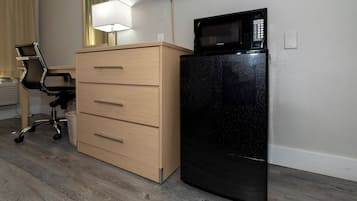 A small room with a wooden dresser, a black refrigerator, a desk with a chair, and a mirror on the wall.