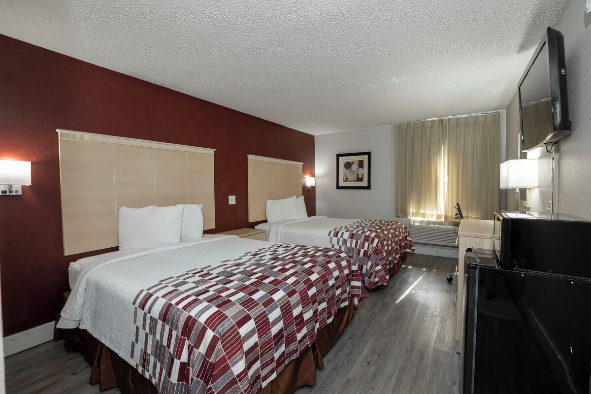 A hotel room with two beds, a television, a desk, and a window with curtains.